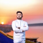 A man in white casual attire standing at sunset by a colorful boat and calm sea.